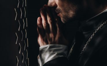 cropped view of catholic priest praying near confessional grille in dark with rays of light